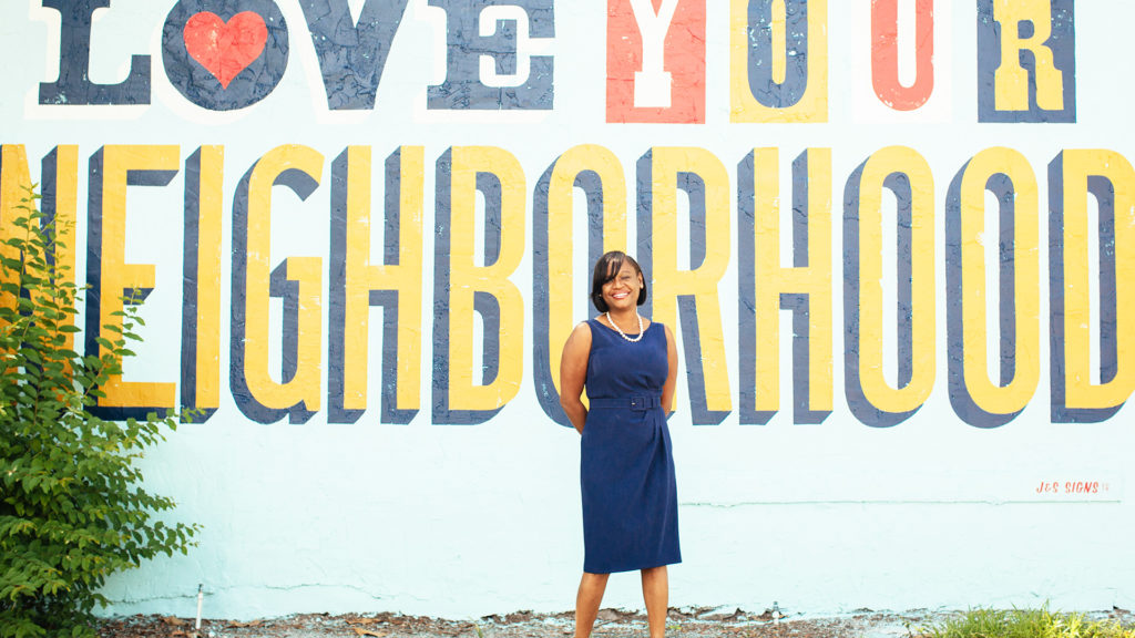 Deputy Mayor Kanika Tomalin stands in front of a blue wall with the words Love Your Neighborhood painted on it.
