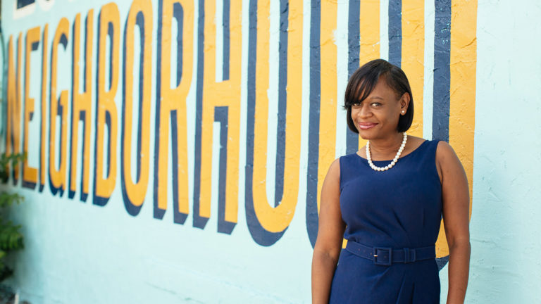 Deputy Mayor Kanika Tomalin stands in front of a blue wall with the word "neighborhood" written in yellow.