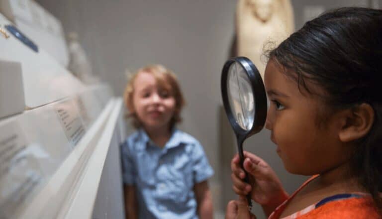 A young child looks through a magnifying glass at art as another child looks at them