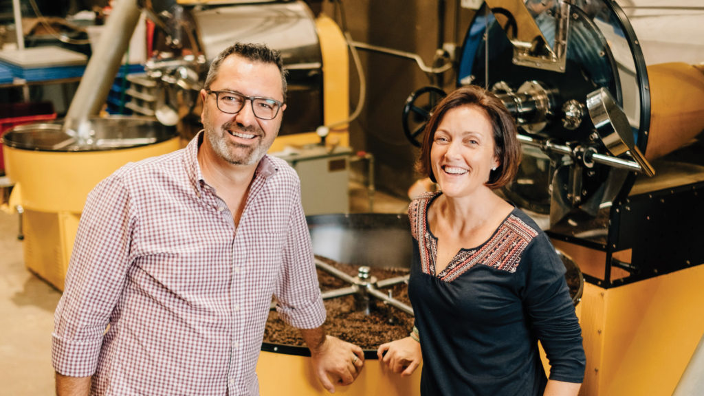 two people smile in front of a large coffee roaster
