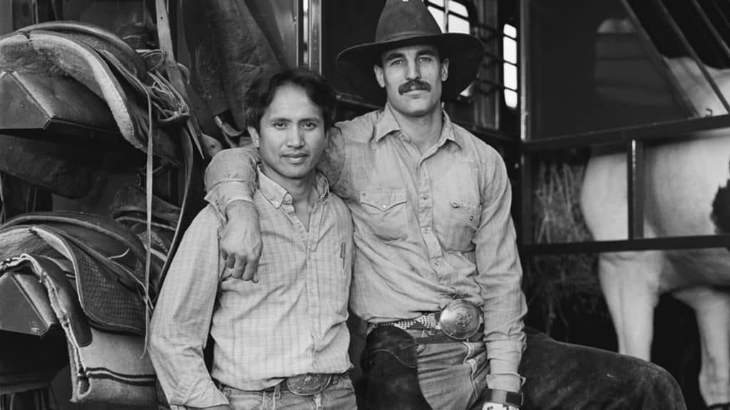 A black and white photograph of two cowboys. The one on the right is wearing a cowboy hat and has his arm around the shoulder of the other cowboy.