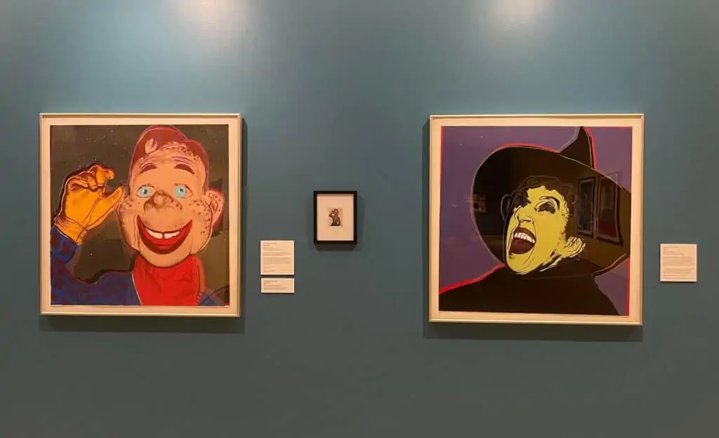 Two pop art photos in a gallery. Oe photo features a green witch laughing in front of a purple background. The right photo features a western style dummy smiling