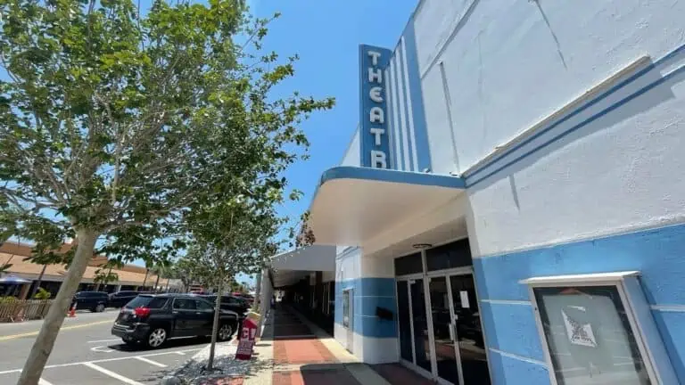 Exterior of a movie palace with a tall blue marquee, and a white facade. An empty ticket counter is visible.