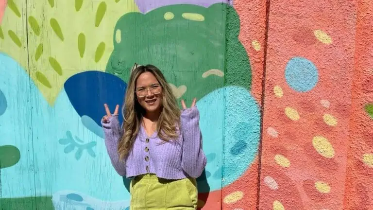 A person in a purple sweater and a green skirt standing in front of rainbow colored mural