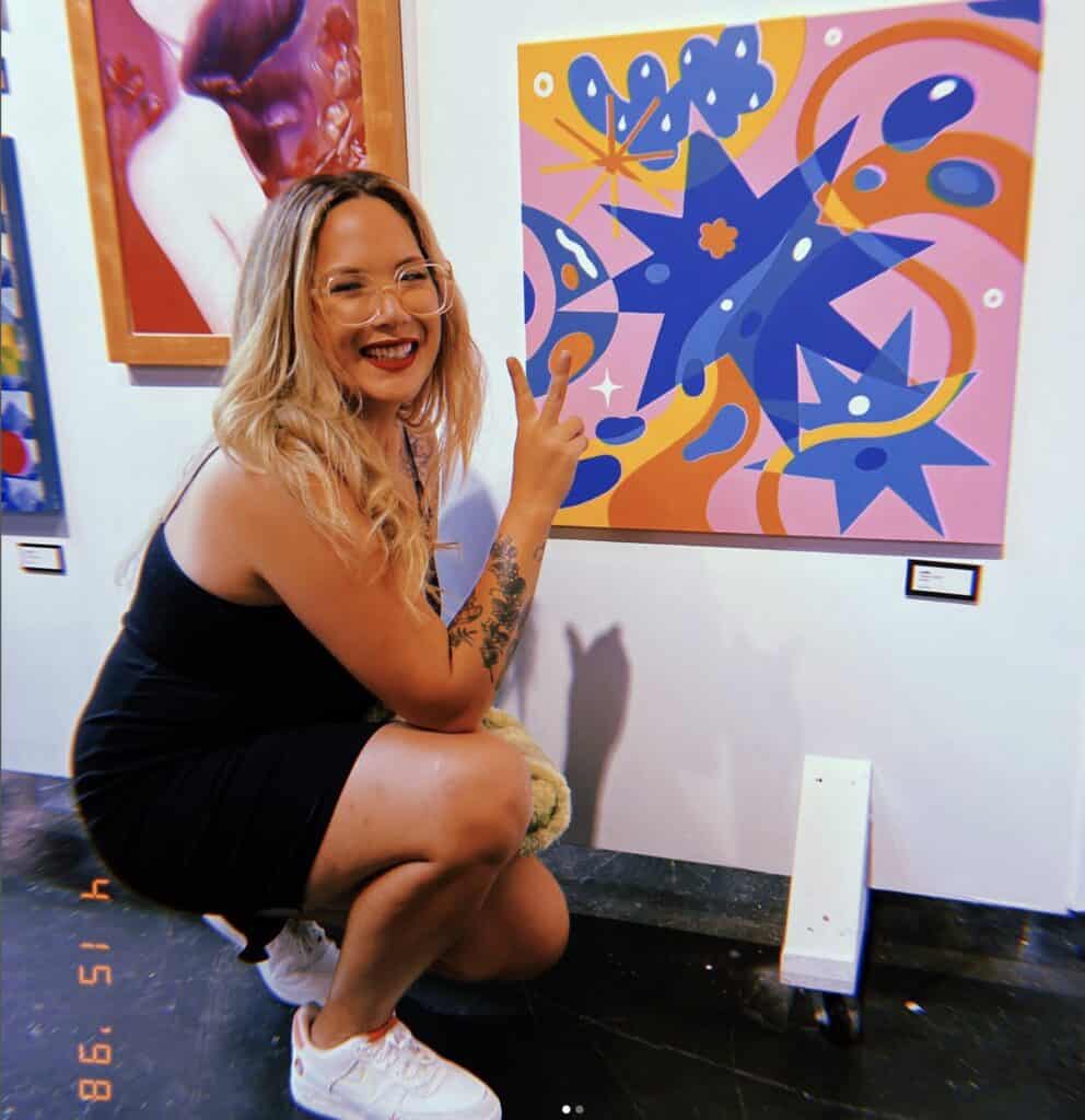 An artist in a black dress, wearing glasses, poses in front of a painting with purple and blue shapes painted on a light pink canvas. 