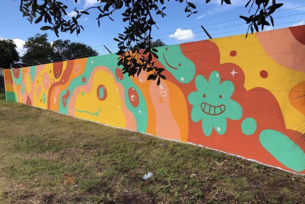A long red, orange, blue and pink mural featuring smiling clouds with tree branches hanging over the front of it.