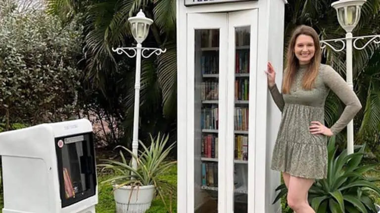 A tall telephone booth repurposed into a library. A person poses in front of the telephone booth.