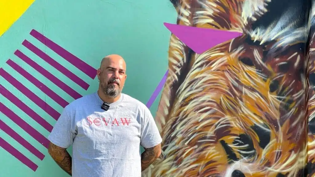 artist stands infant of their mural. The background is teal with a cheetah head painted on the right side. A geometric sequence of purple lines forms a diamond in the right hand corner.