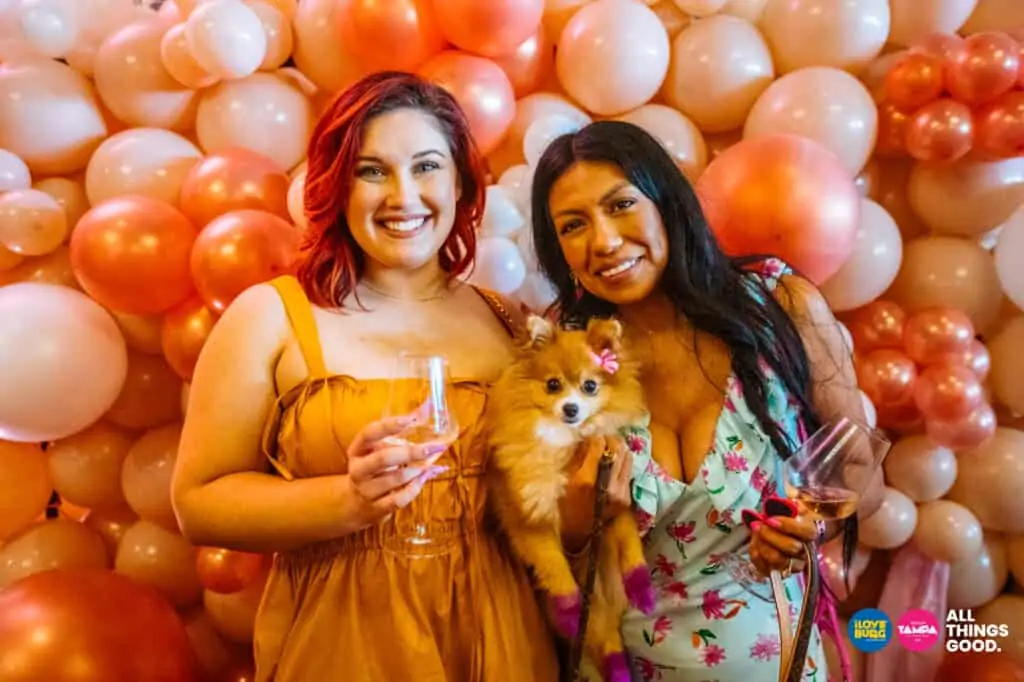 two friends pose with their pomeranian in front of a pink and rose balloon wall