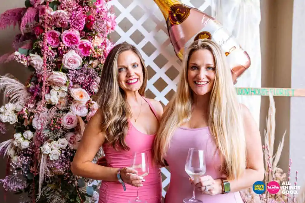 two friend pose with wine in front of a flower wall