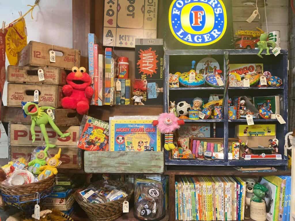 An assembly of vintage toys and books for children arranged on wooden shelves. Kermit the Frog and Elmo are set up in the corner.