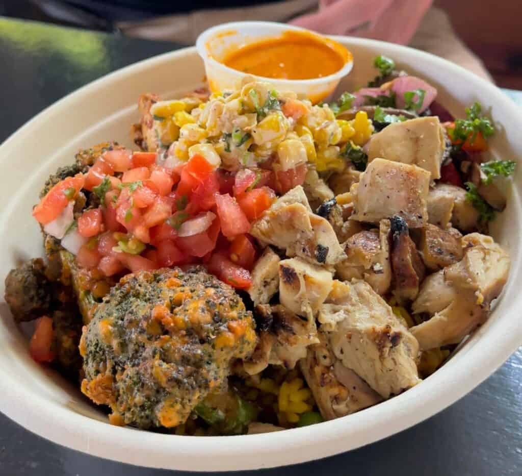 a bowl featuring broccoli covered in hot sauce, seasoned chicken, diced tomatoes, and corn salad. 