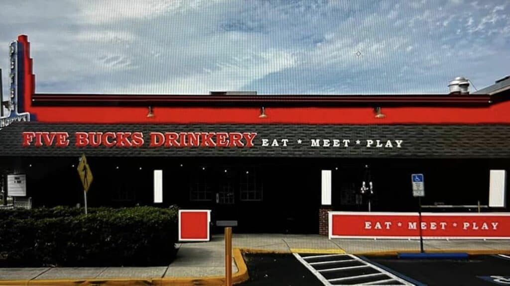 Exterior of a bar with a red trim awning. Covered outdoor seats are shown next to the parking lot. A sign reading "eat drink play" is visible in the foreground.