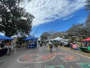 A street festival with vendors. There's a mural painted on the street. Green trees on either side of the road.