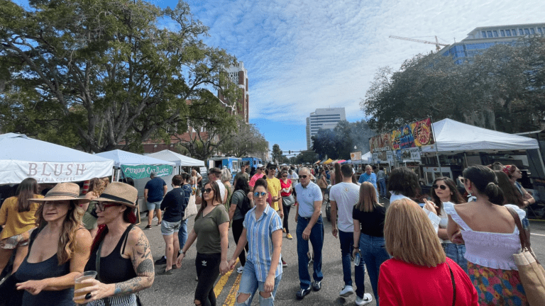 people gather out in the streets during a large vendor festival. Big green trees frame the photo and the sky is deep blue.