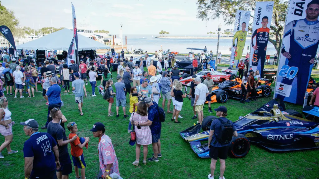 A party at a waterfront park. race cars are parked in the grass as attendees gather around them.