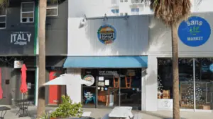 Exterior of a business with a white exterior, a blue awning and a blue circular sign above the door. Two tables with umbrellas are outside.