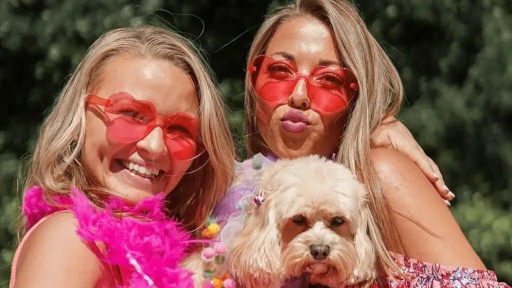 Two women dressed in pink in a car with a dog. they each have on a pair of heart shaped sunglasses