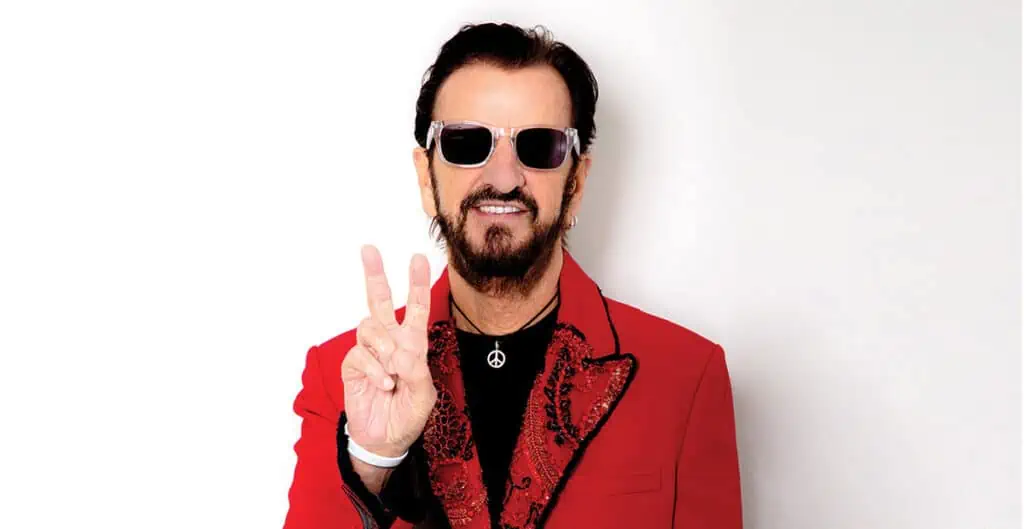 Ringo Starr wearing a red suit and sunglasses holds up a peace sign