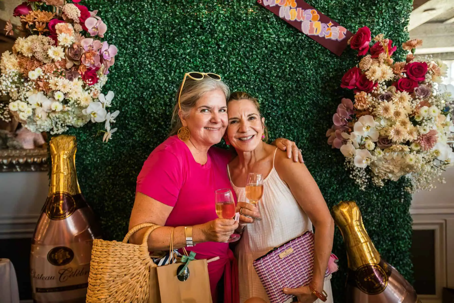 two friends smile with their wine glasses in front of a green and pink floral wall