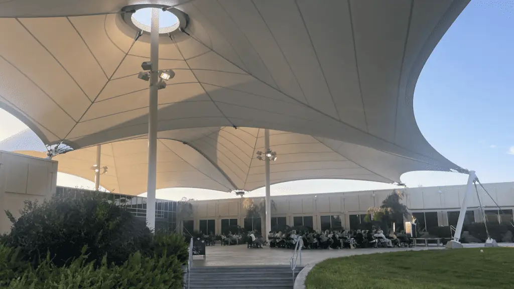 a group of people listen to an author reading under a large shade structure on a stone patio