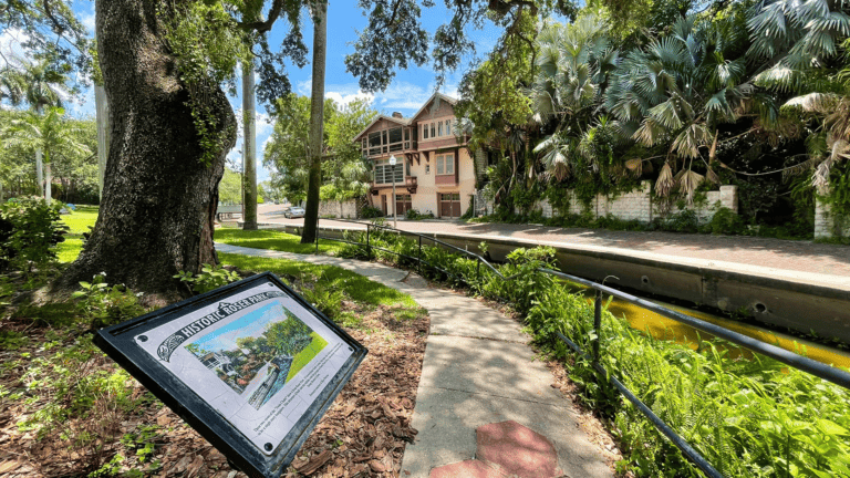 A welcome sign to a historic neighborhood with brick streets, a creek, and tall shade trees