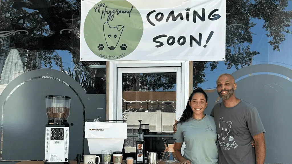 Two people standing side by side outside a bar with a coffee cart to their left. A coming soon banner with a green logo featuring a cartoon dog is on the left