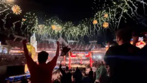 fireworks go off at an outdoor stadium, a wrestling ring is lit up in the center of the stadium