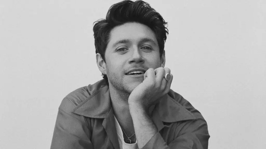 Niall Horan poses resting his chin on his hand, elbow on a table in black and white