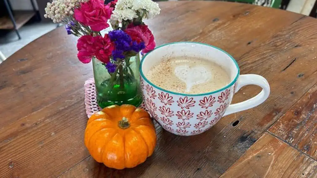 a mug with pumpkin spice latte sits on a wooden table with a vase of flowers and a small pumpkin