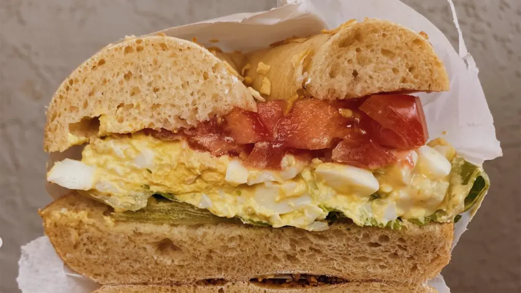 a toasted bagel sandwich loaded with eggs, tomatoes and fresh greens