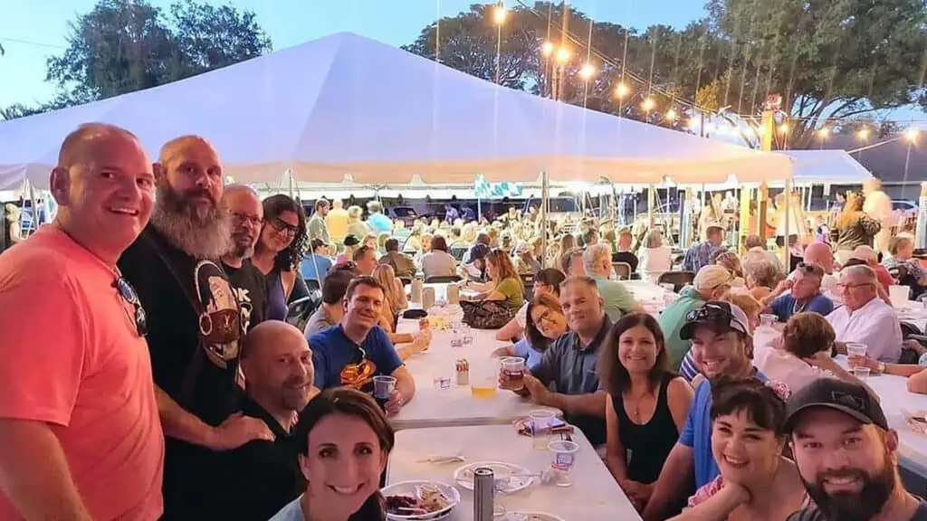 attendees enjoying an outdoor festival at a long picnic table. A large white tent is in the background