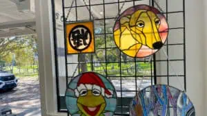 several stained glass art pieces hanging on a wire stand including a kermit the frog and a greyhound portrait