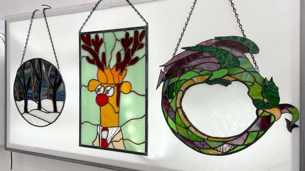 three stained glass art pieces hanging on a lightbox, one is a muppet, one is a dragon eating its tail, and one is of trees in snow