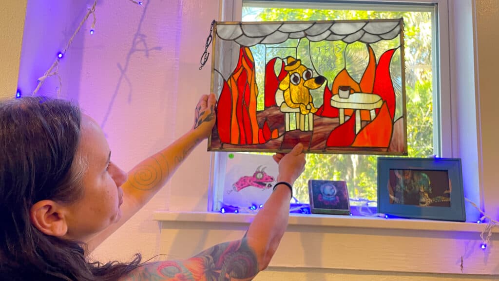 Artist Jodie Chemes holds up a stained glass art piece of the "this is fine" meme with a dog in a burning room