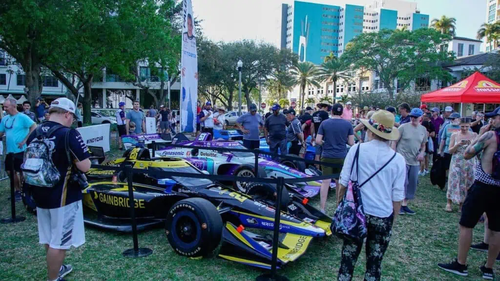 An Indycar is displayed at the center of a park with multiple people gathered around it. Vendor tents are visible in the background