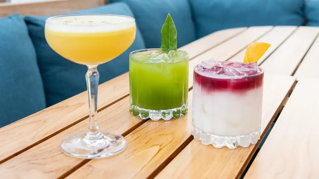 three bright cocktails in glasses on a wooden table. One cocktail is bright yellow, the middle one is green, and the cocktail on the right is white with a purple/pink top