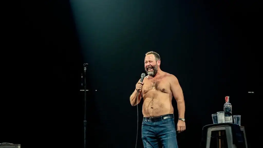 a comedian, shirtless, on stage with a spotlight on.