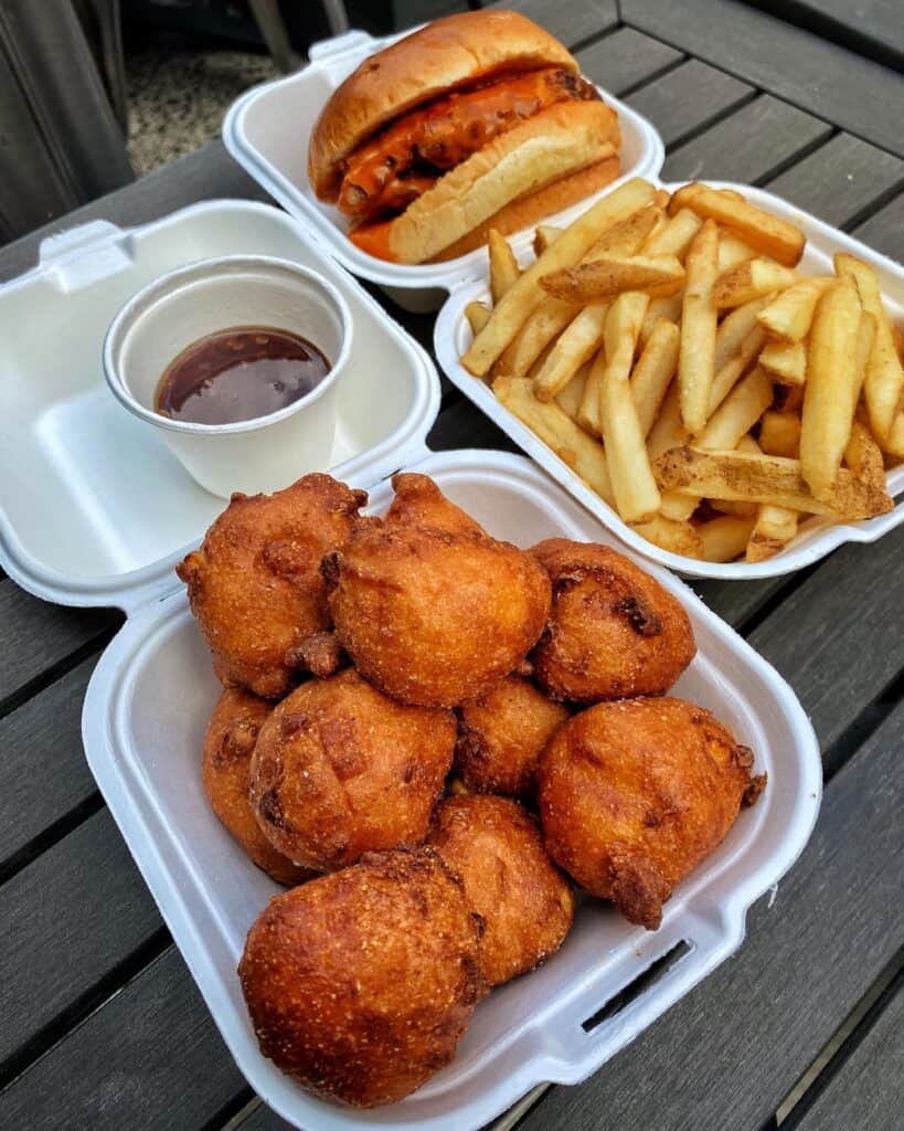 A serving of hush puppies and fries in two containers with a chicken sandwich. 