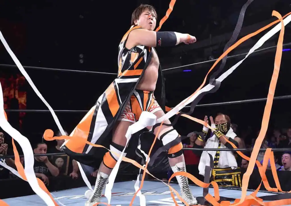 a performer in orange and black trunks strolls through a ring covered in ribbons