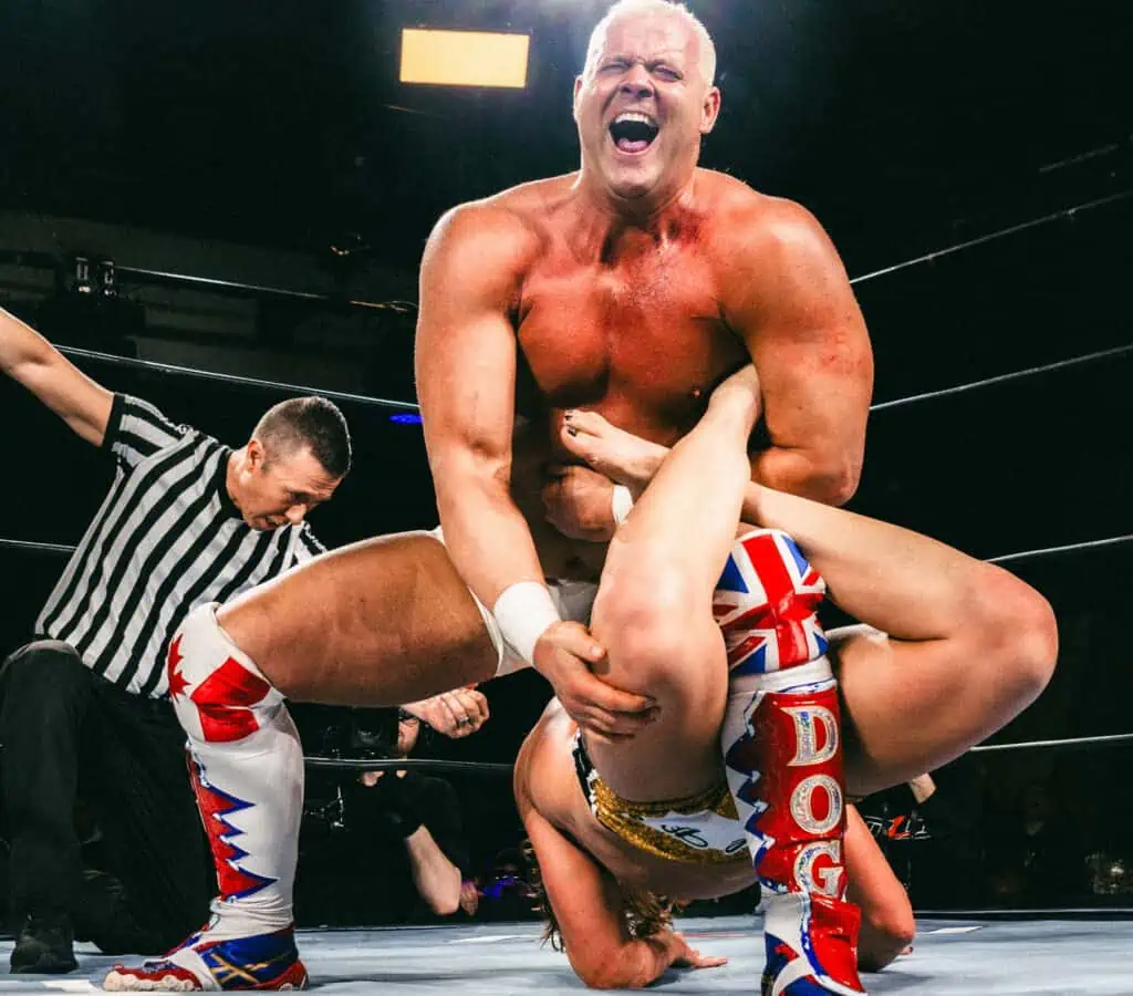 a performer in blue and red trunks performs a leg submission hold in the middle of a wrestling ring