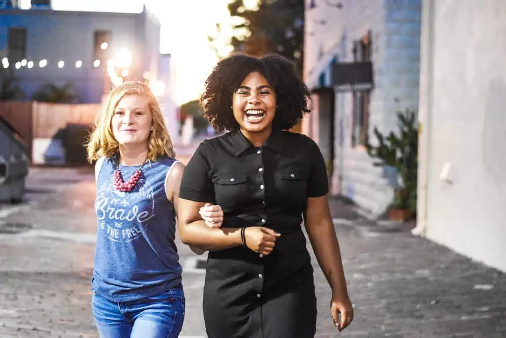 Two girls, one white and one African American holding arms, walking and laughing. 