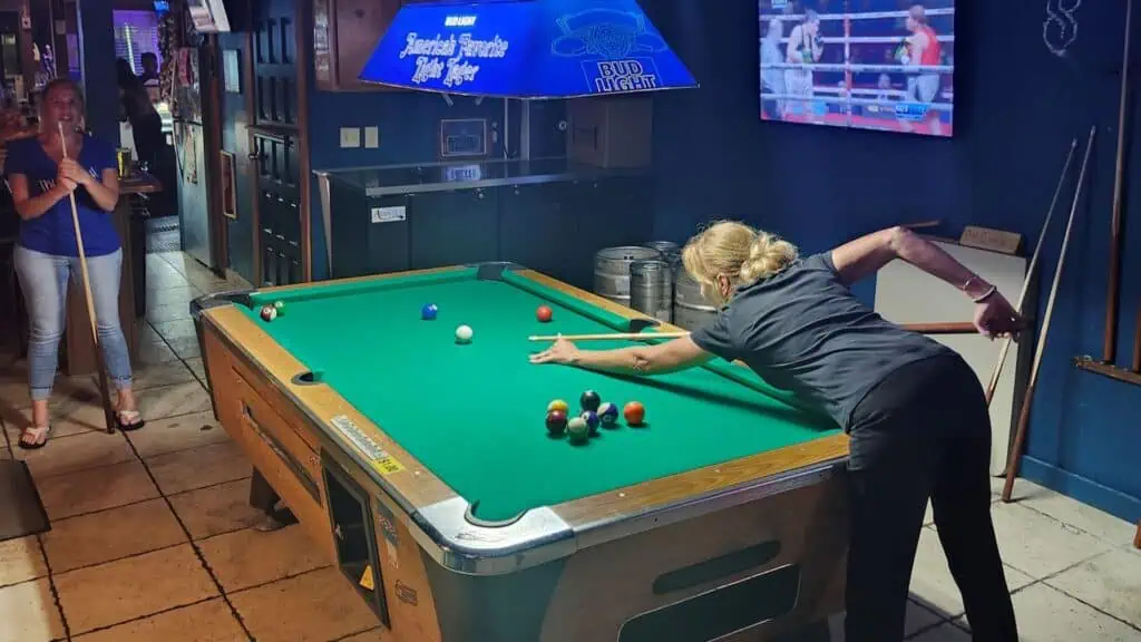 2 patrons play pool at the bar, one of them is leaning forward to hit her shot