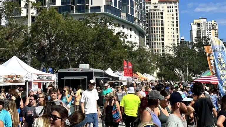 a gathering f people enjoy shopping at the outdoor farmers market in St. Pete, Florida. Food trucks and vendor tents with flags flying above the crowd are visible