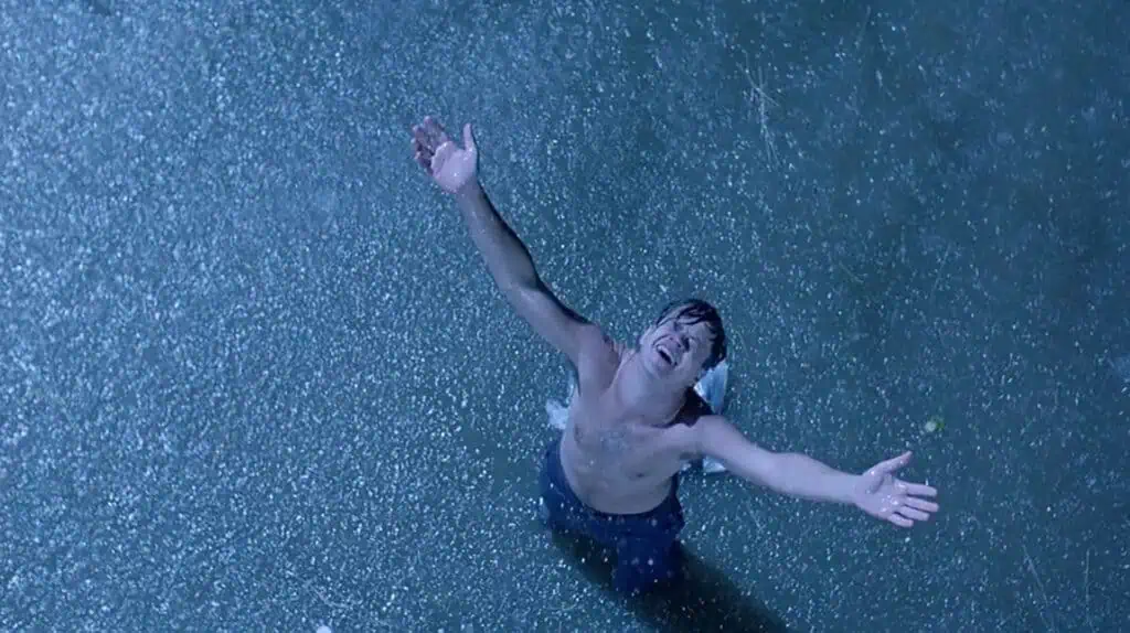 a man stands shirtless in the rain with his arms outstretched to the sky 