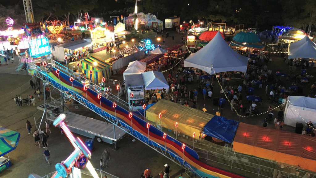 acolorful slide is in the foreground. The photo showcases the entirety of a small neighborhood carnival with vendor tents and rides