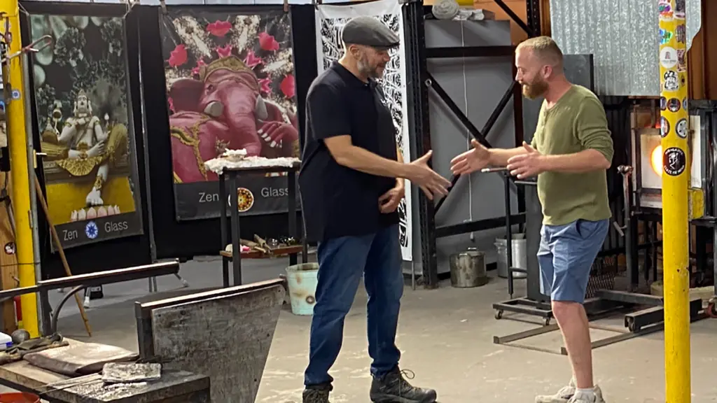 two people stand at the center of a glassblowing studio during a TV taping