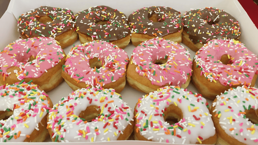 assorted frosted donuts in a box. Donuts are arranged with pink frosted, chocolate frosted, and vanilla frosted. 