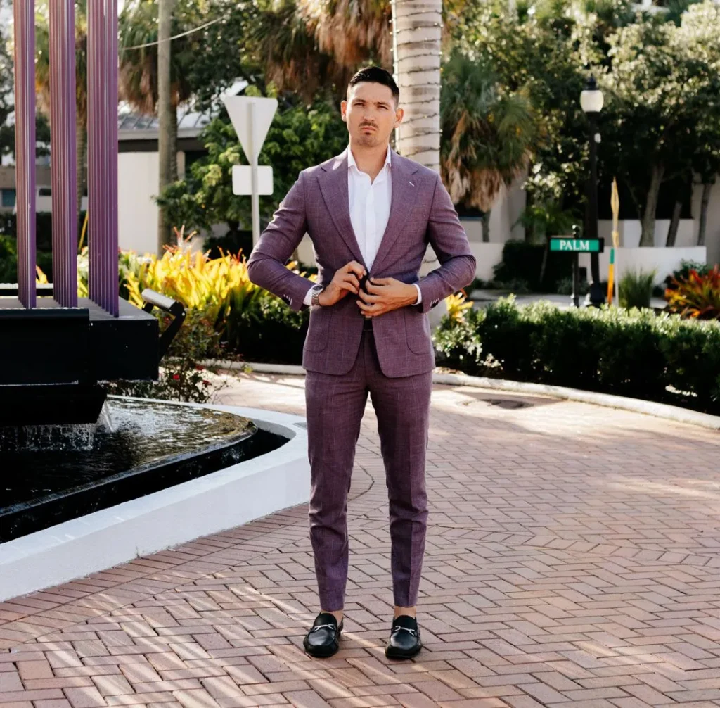 Man wearing purple Tweeds suit with white shirt. Standing on brick pathway with plants and vegetation surrounding in the background