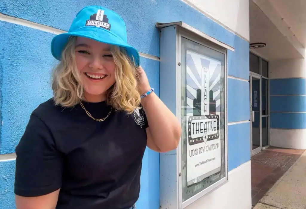 girl wearing bur hat smiling in front of theatre poster.